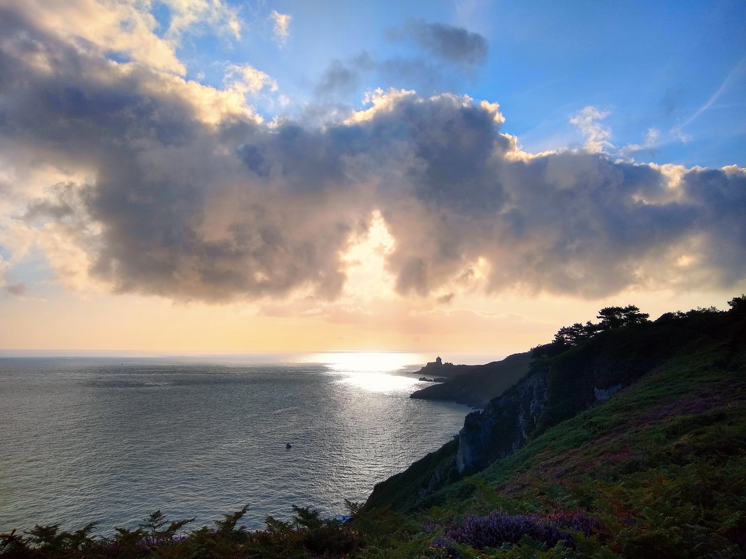 Falaises et végétation côtière devant la mer, avec le soleil qui traverse de grands nuages.