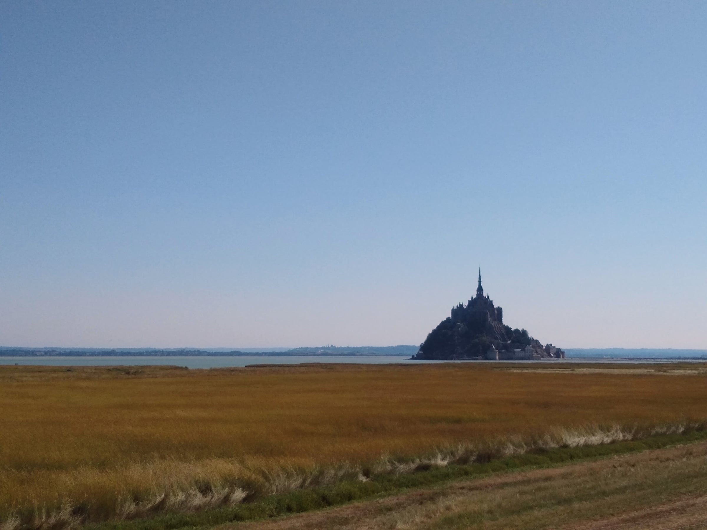 Mont-Saint-Michel au loin, derrière une vaste étendue d’herbes et de sable sous un ciel bleu.