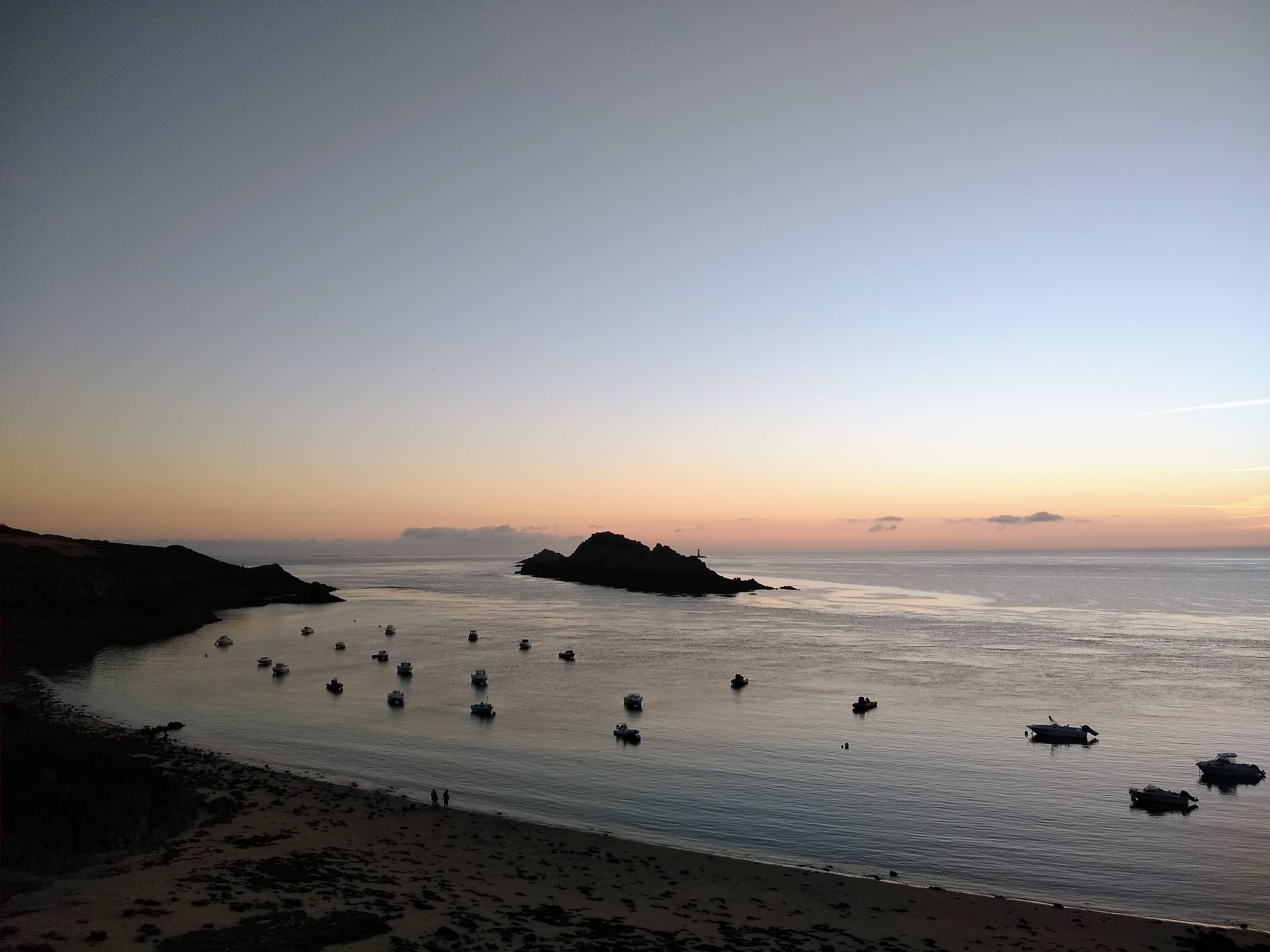 Bateaux au mouillage entre la plage et des îlots, sous un ciel aux teintes orangées.