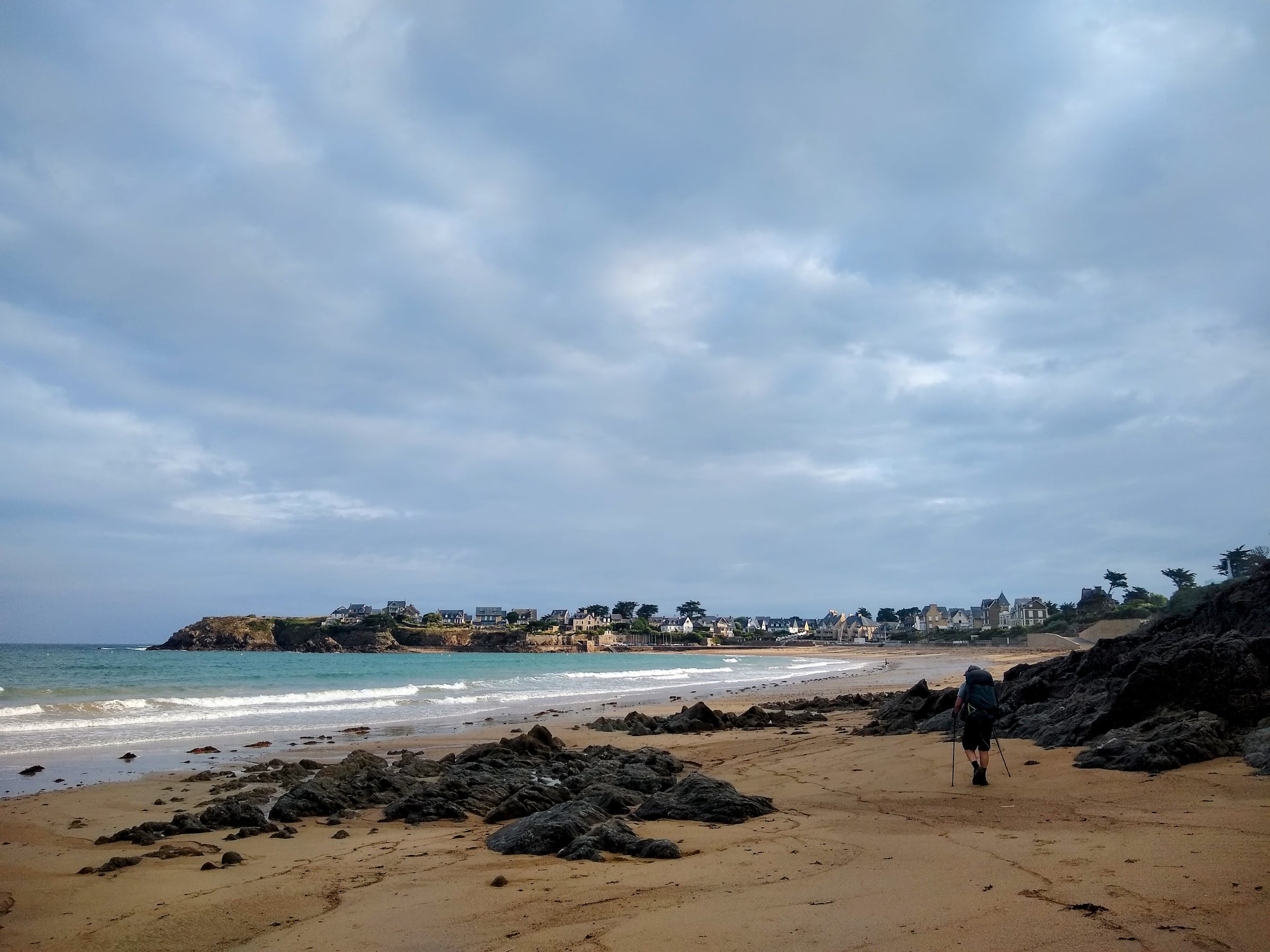 Randonneur sur une plage bordée de rochers, devant la mer et des maisons au loin.