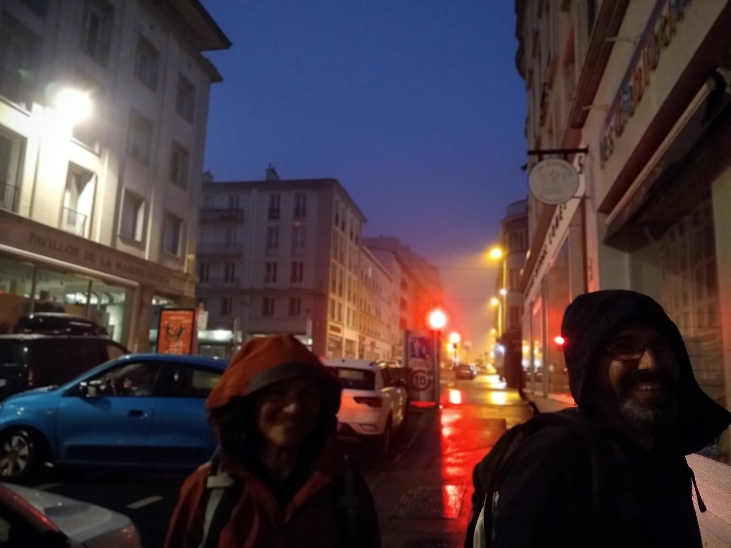 Deux personnes en vestes à capuche dans une rue mouillée et éclairée, à la nuit tombée.