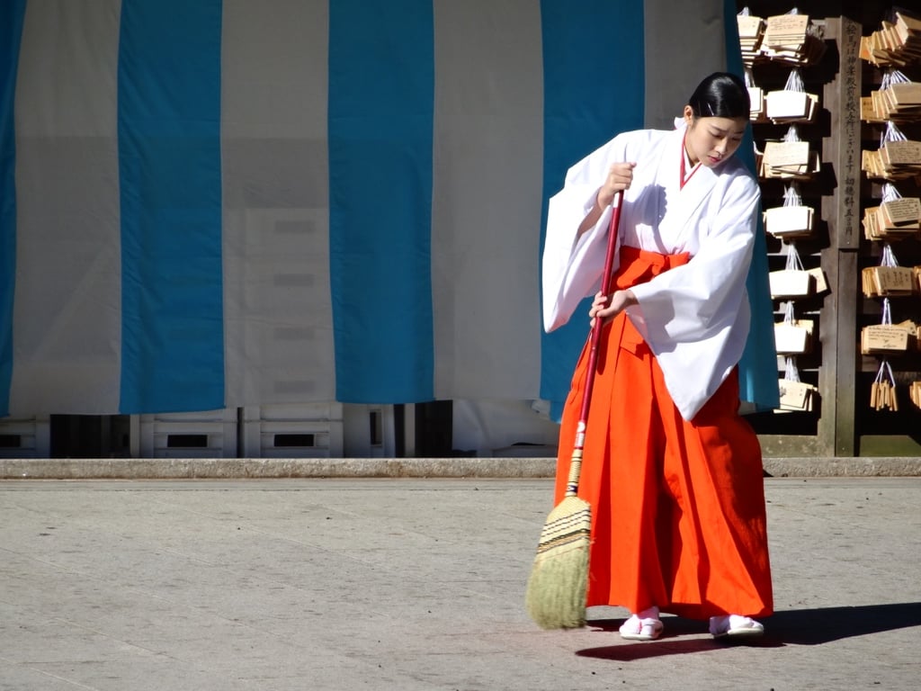 Femme en tenue blanche et rouge balayant devant un bâtiment de sanctuaire.