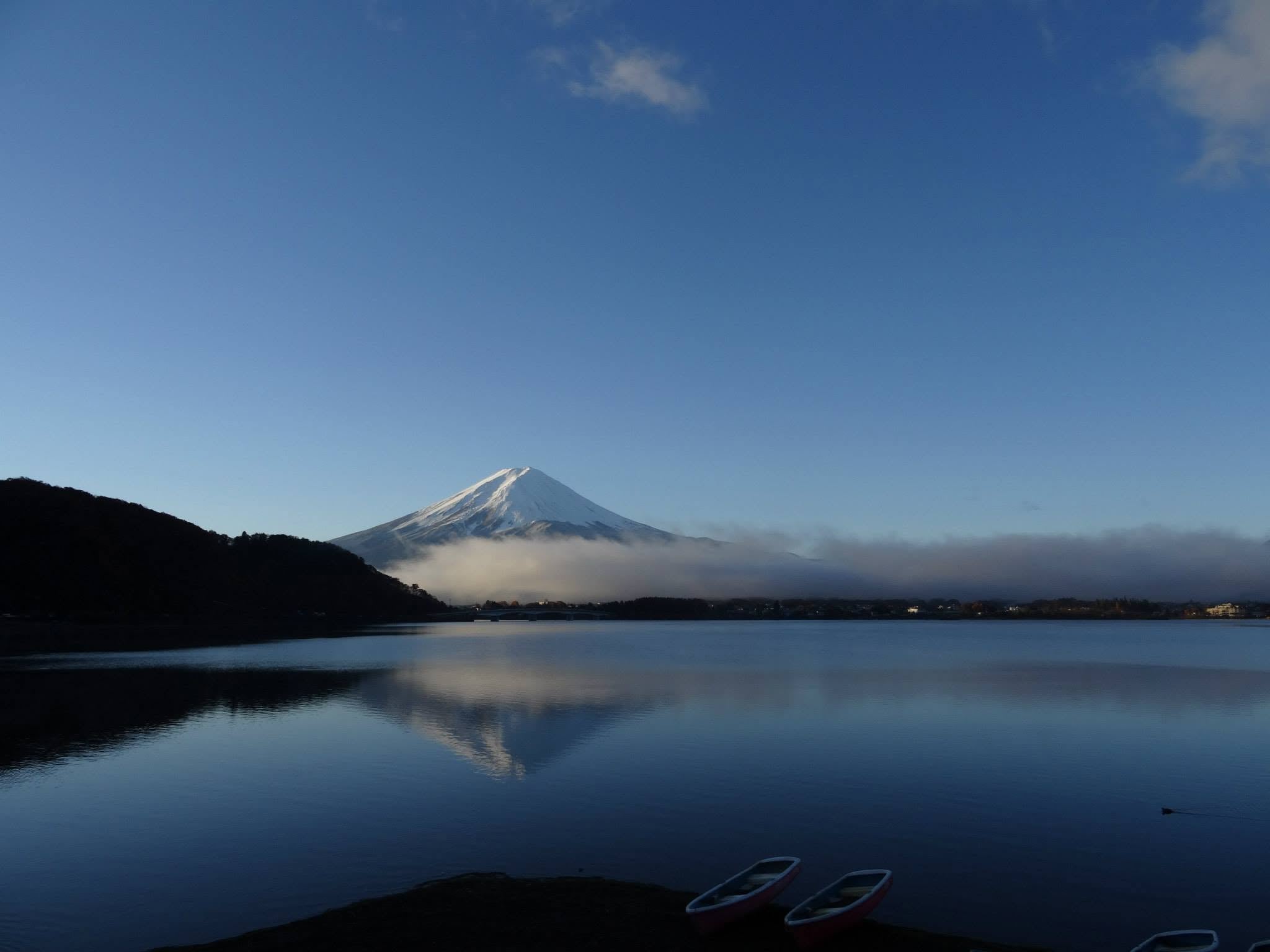 Mont Fuji et son reflet dans un lac calme, avec des nuages bas et des barques au premier plan.