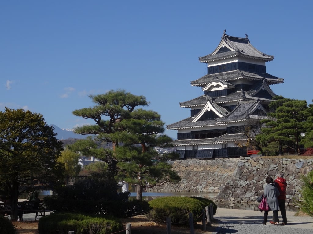 Château japonais noir et blanc derrière des pins, des murs de pierre et quelques visiteurs.