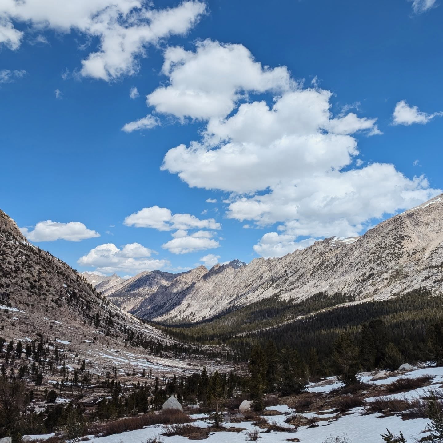 Vallée de granit bordée de pins, avec des plaques de neige et des nuages au-dessus des sommets.