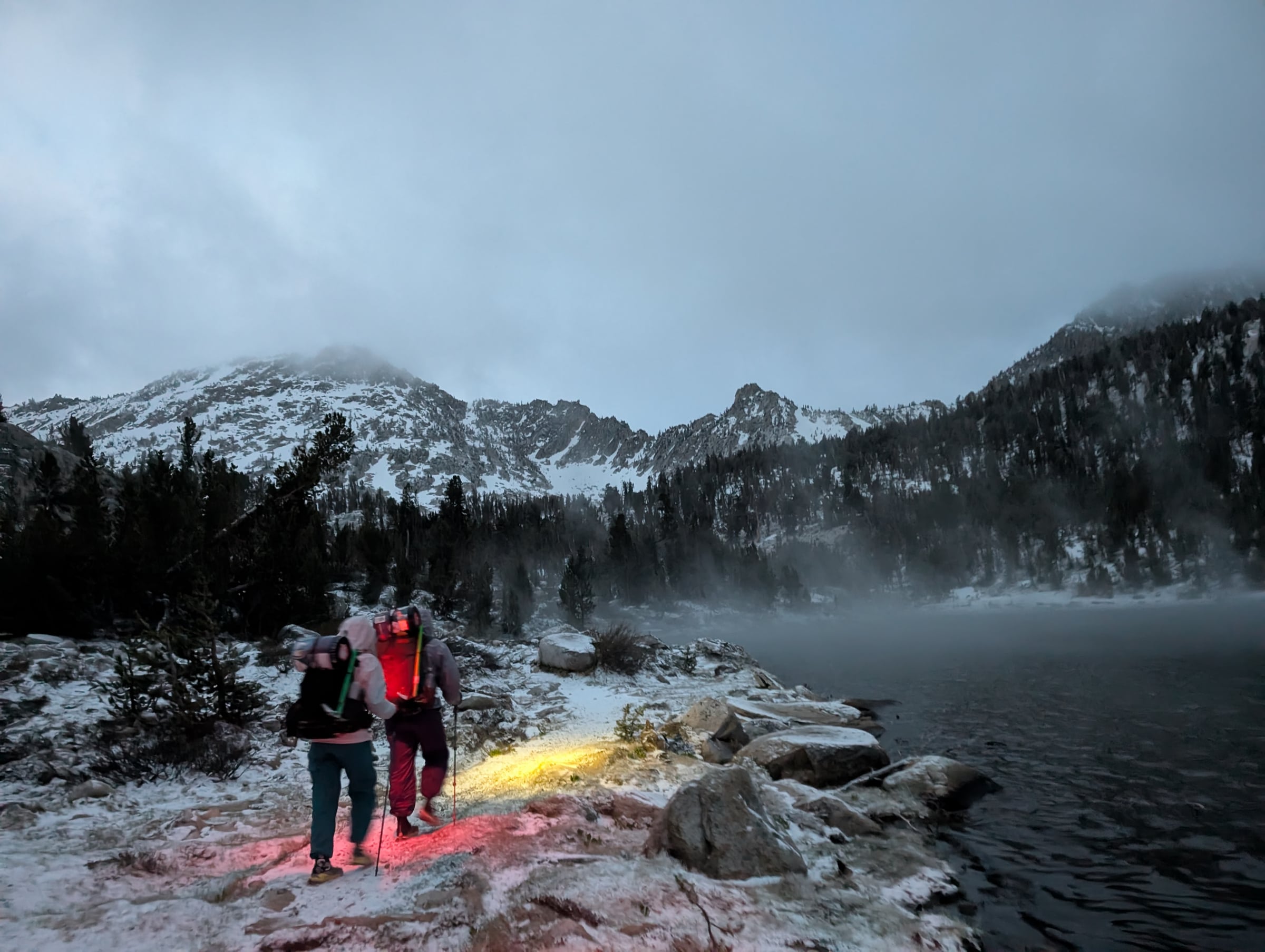 Deux randonneurs avancent à la lumière de leurs frontales au bord d’un lac enneigé dans la brume.