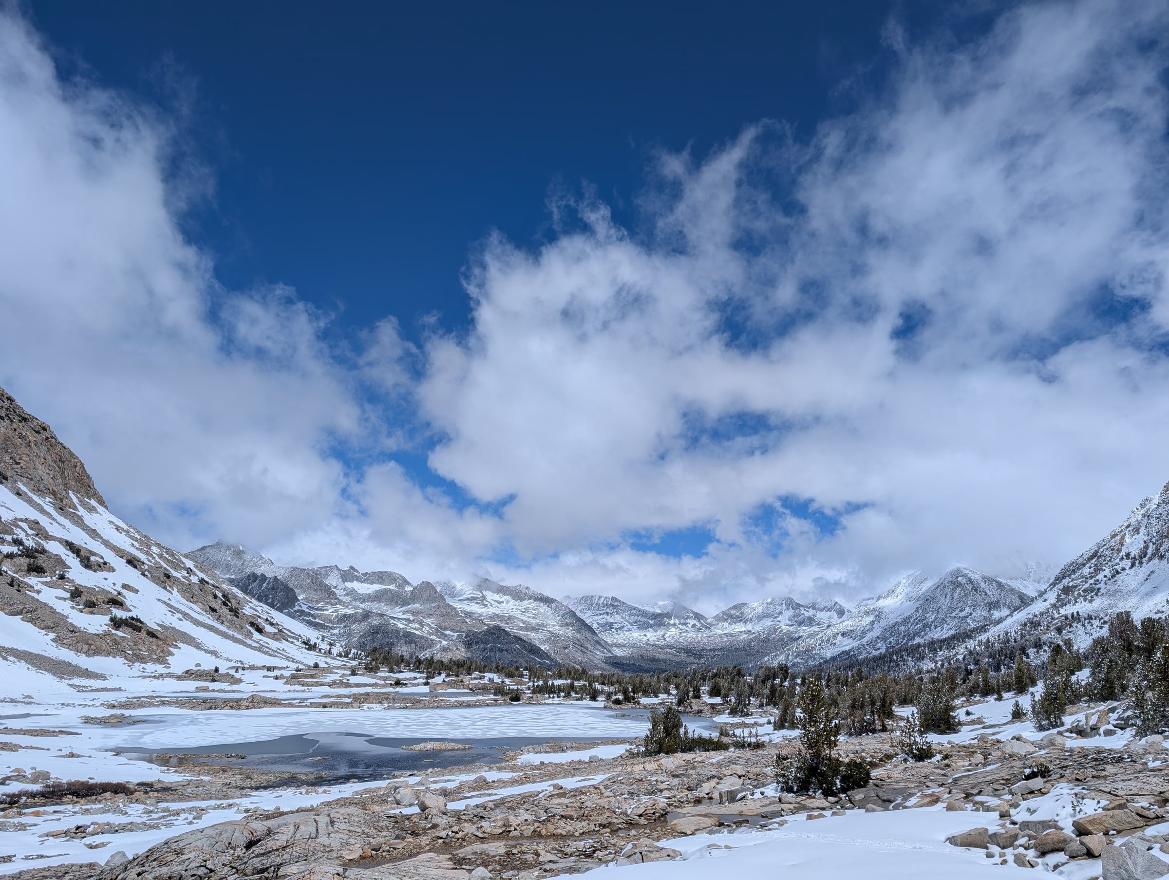 Lac partiellement gelé entouré de rochers, de pins et de sommets enneigés.