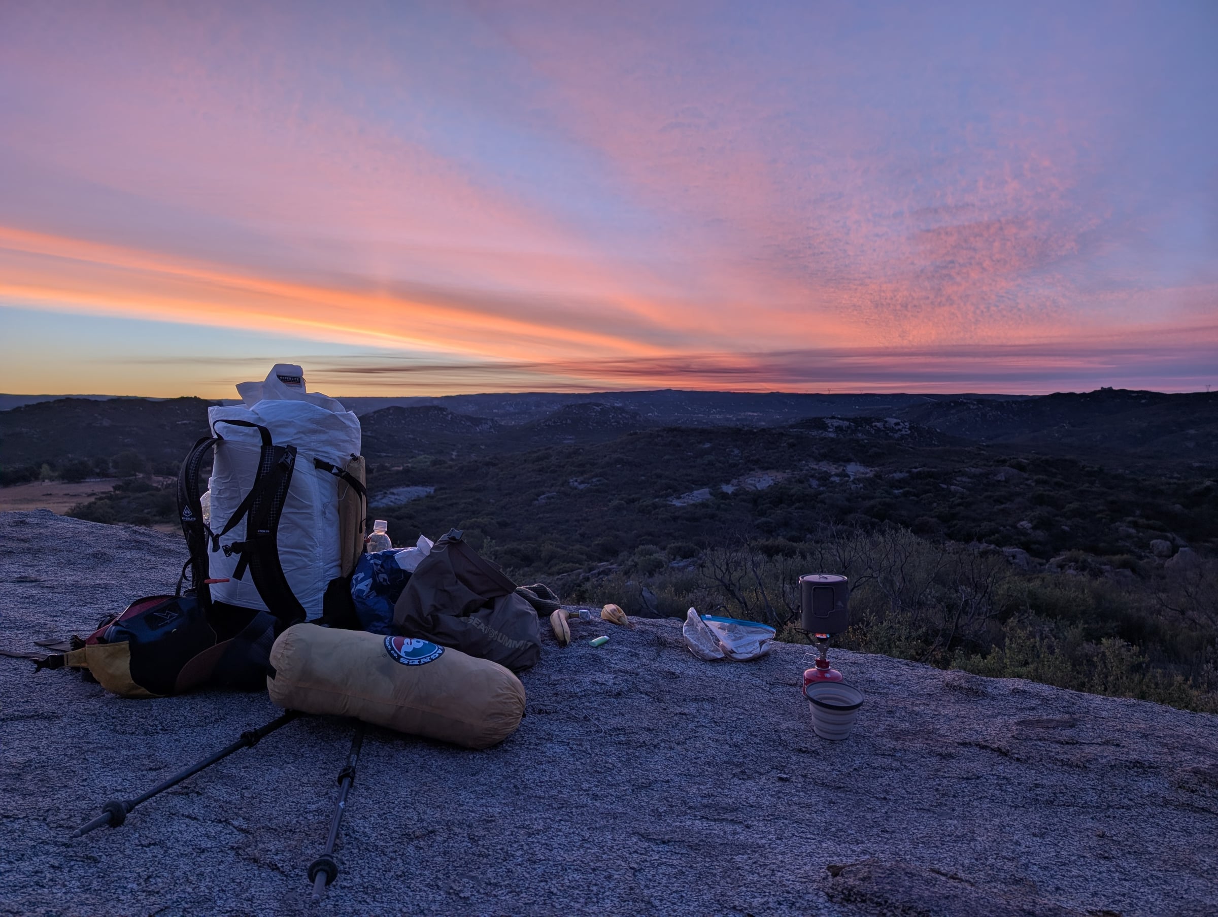 Sac à dos, réchaud et équipement de bivouac sur un rocher, devant un ciel rose et orangé.