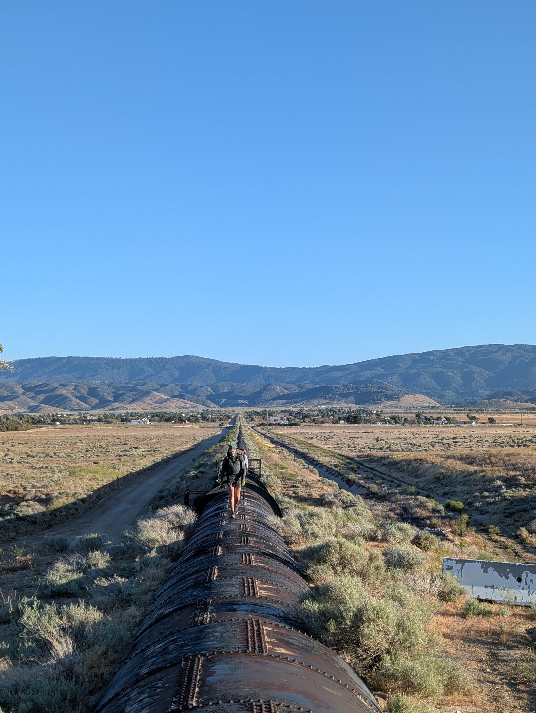 Randonneur sur un long conduit métallique qui traverse une plaine sèche vers les montagnes.