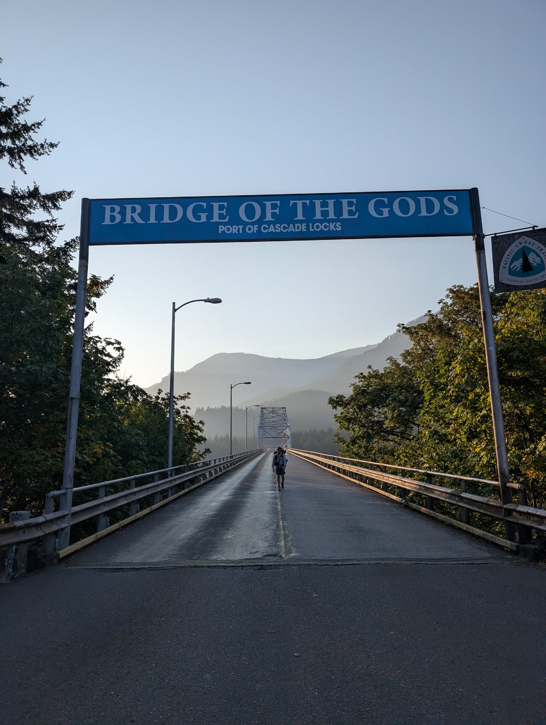 Randonneur sous le panneau Bridge of the Gods, sur un pont entouré de montagnes boisées.