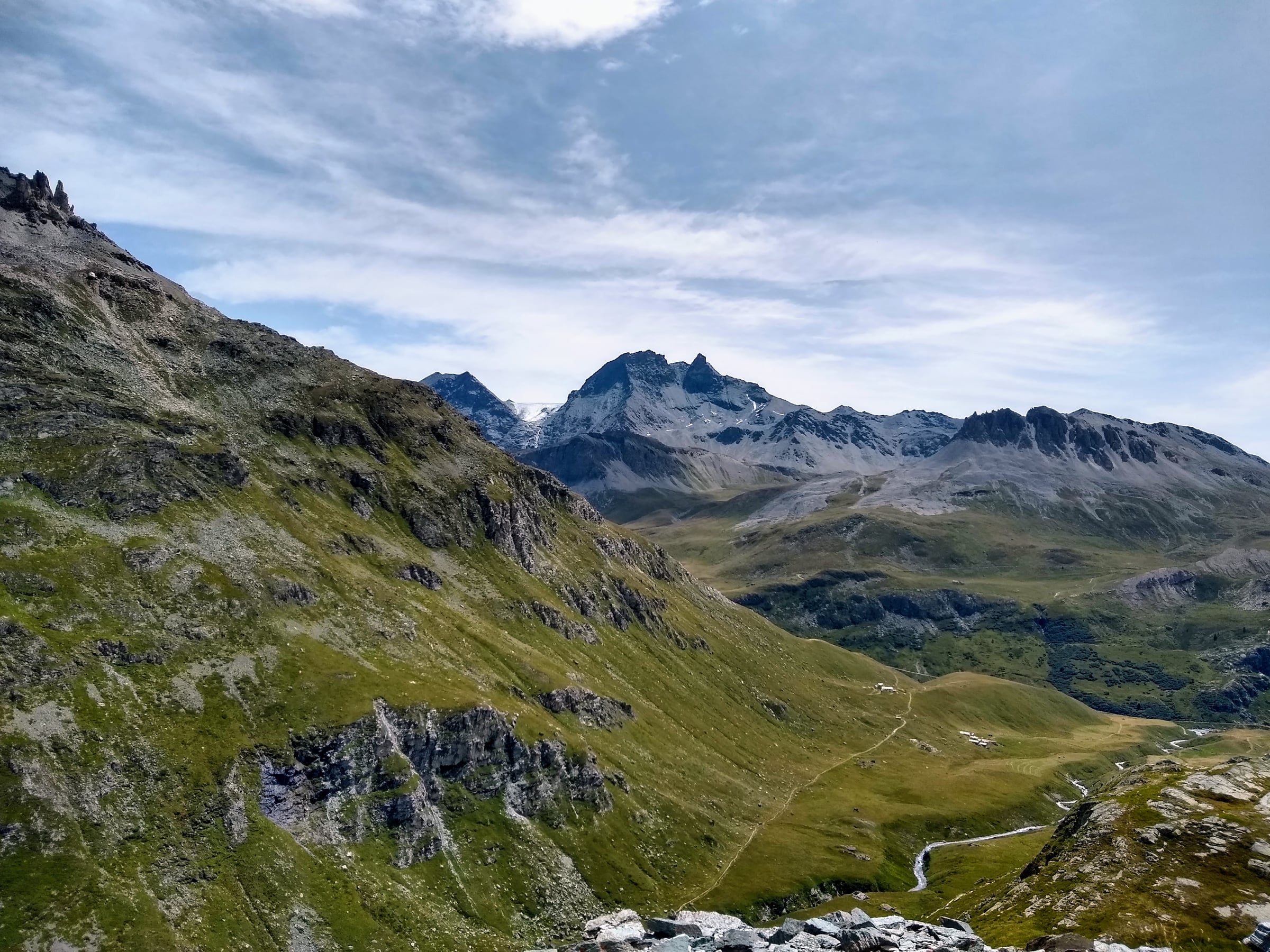 Vaste vallon verdoyant parcouru d’un sentier, sous des sommets rocheux et un ciel nuageux.