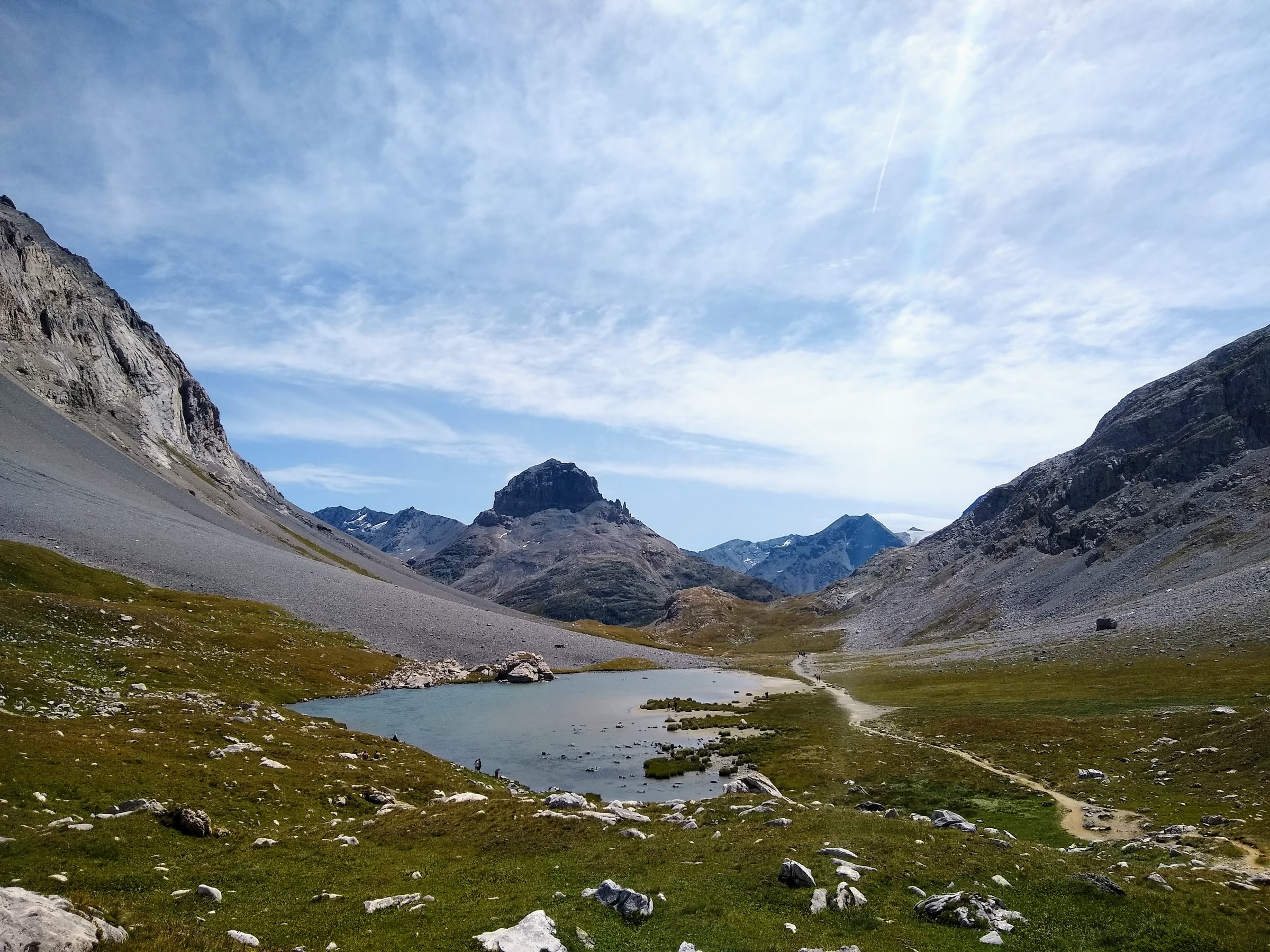 Lac d’altitude au milieu de prairies et de pentes rocheuses, avec un sentier qui longe la rive.